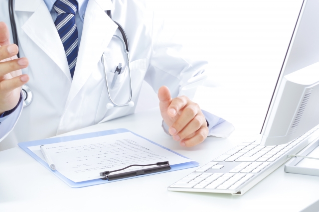 白衣の医師が机でパソコンと書類に向かう