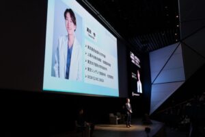 ステージ上の講演者とスクリーンに映る講演者のプロフィール